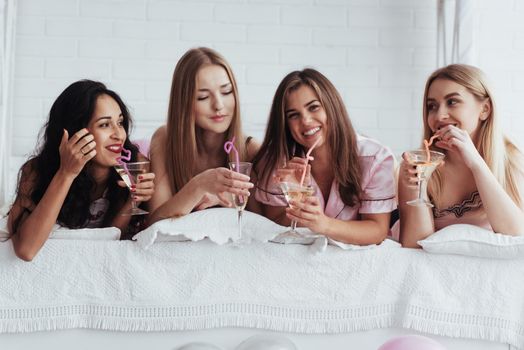 Drinking some good cocktails. Joyful girls in the nightwear lying on the bed in white room and have celebration.