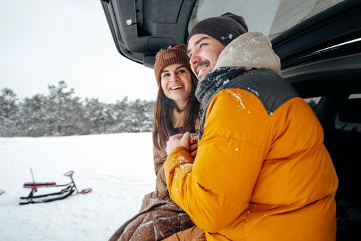 Lovely smiling couple sitting in car trunk in winter forest, close up