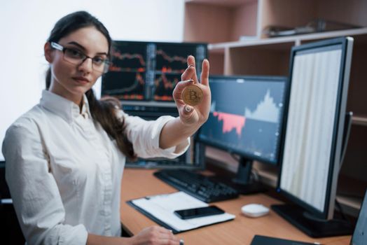 Young freelancer in eyewear. Beautiful woman holding bitcoin in hands while sitting in modern office with many monitors with graphs.
