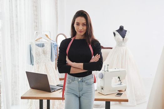 Looking into the camera with arms crossed. Portrait of female fashion designer standing in the workshop and table with instruments behind.