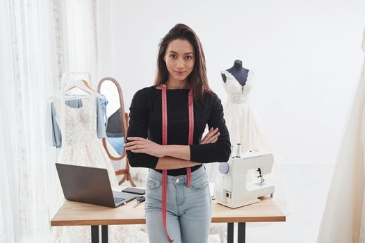 Conception of the wedding clothes. Portrait of female fashion designer standing in the workshop and table with instruments behind.