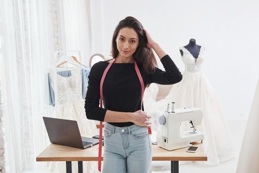 Touches hair. Portrait of female fashion designer standing in the workshop and table with instruments behind.
