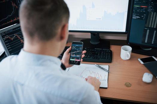 Financial report on the one big graph n the smartphone. Man working online in the office with multiple computer screens in index charts.