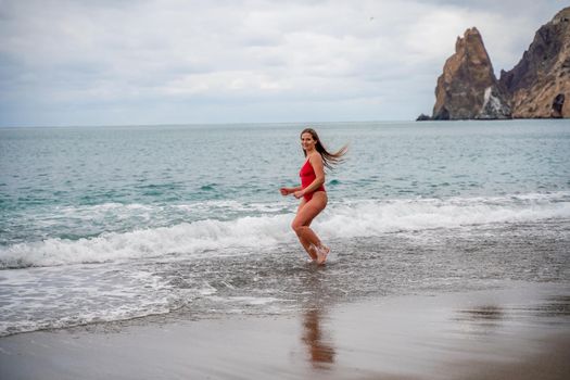 A beautiful and sexy brunette in a red swimsuit on a pebble beach, Running along the shore in the foam of the waves.