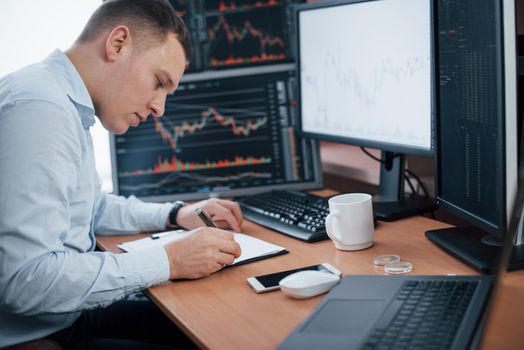 Writing on the notepad. Man working online in the office with multiple computer screens in index charts.