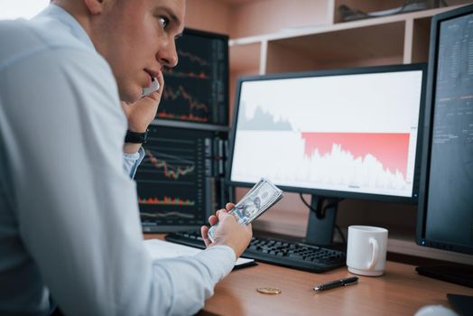 Haves conversation while holding money in the hand. Man working online in the office with multiple computer screens in index charts.