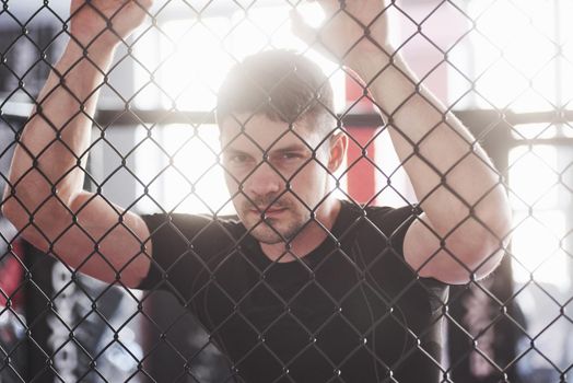 Close up view. Sportive guy in black shirt stands behind the fence in the room with daylight behind.