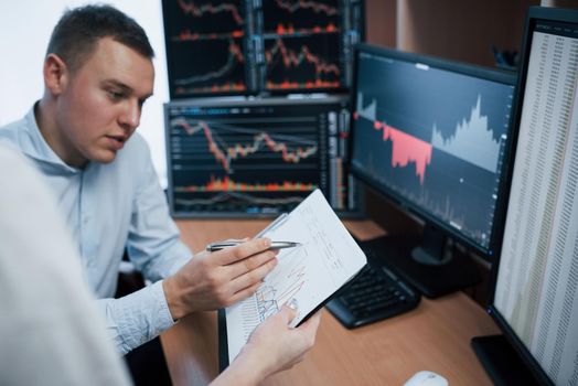 Notepad in hands with some graphs. Team of stockbrokers are having a conversation in a office with multiple display screens.