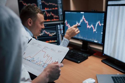 Girl holds paper with information. Team of stockbrokers are having a conversation in a office with multiple display screens.