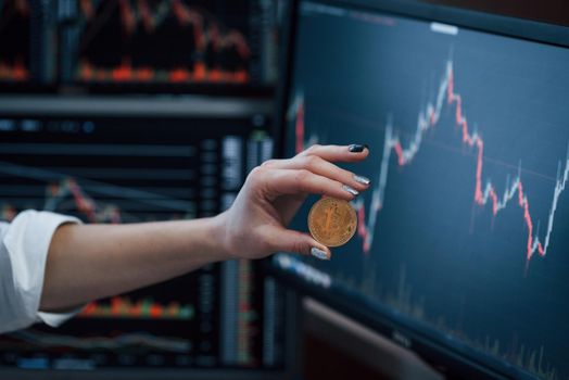 Close up view. Woman's hand holding bitcoin in modern office with many monitors with graphs.