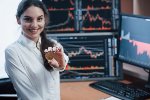 Nice daylight. Beautiful woman holding bitcoin in hands while standing in modern office with many monitors with graphs.