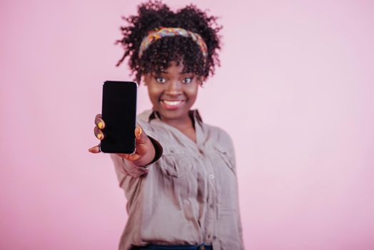 Smiling and feeling joy. Holding black phone in hand. Attractive afro american woman in casual clothes at pink background in the studio.