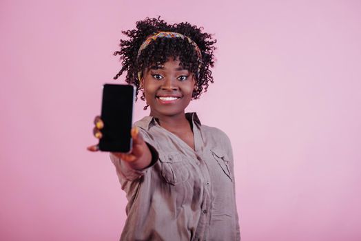 Holding black phone in hand. Attractive afro american woman in casual clothes at pink background in the studio.