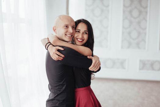 Cute and cheerful people. Hug each other and smiling. Portrait of happy couple indoors. Bald guy and brunette woman stands in the white room.