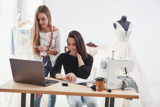 Looking on the laptop. Two female fashion designers works on the new clothes in the workshop.