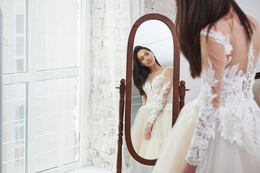 Young bride looks in the mirror. The process of fitting the dress in the studio of hand crafted clothes.