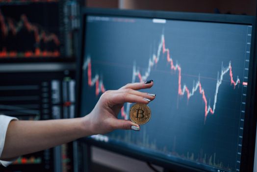 Front view. Woman's hand holding bitcoin in modern office with many monitors with graphs.