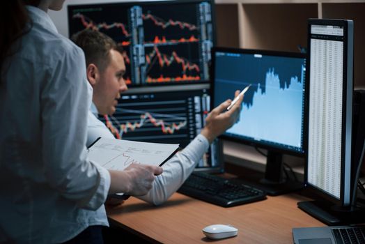 Man shows results. Team of stockbrokers are having a conversation in a office with multiple display screens.