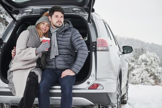 Conception of love and family. Nice couple have weekend at countryside at winter time. Sitting on the rear part of modern car.