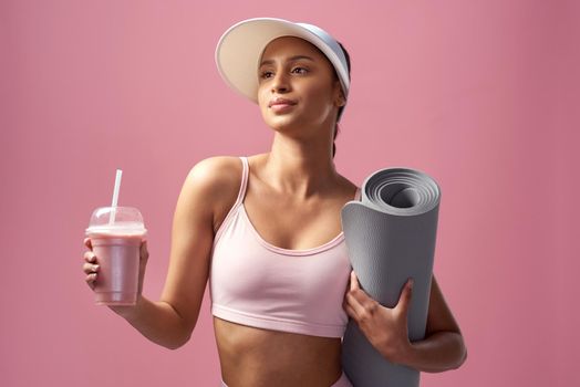 Cropped shot of an attractive and sporty young woman posing with an exercise mat and a smoothie in studio against a pink background.
