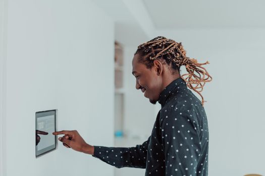 Smiling African American man using modern smart home system, controller on wall, positive young man switching temperature on thermostat or activating security alarm in apartment. High quality photo