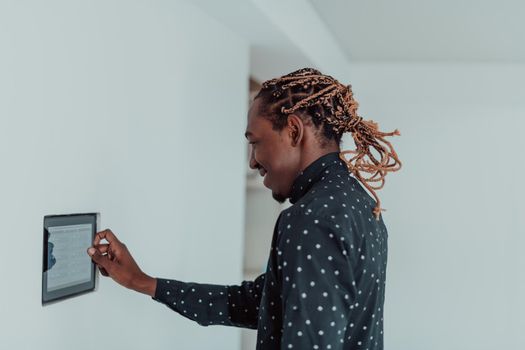 Smiling African American man using modern smart home system, controller on wall, positive young man switching temperature on thermostat or activating security alarm in apartment. High quality photo