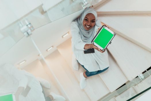 Young African modern Muslim woman using tablet computer while sitting on the stairs at home wearing hijab clothes top view. High-quality photo