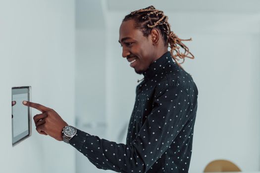 Smiling African American man using modern smart home system, controller on wall, positive young man switching temperature on thermostat or activating security alarm in apartment. High quality photo