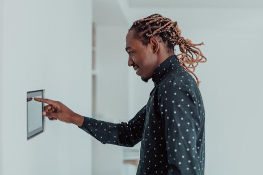 Smiling African American man using modern smart home system, controller on wall, positive young man switching temperature on thermostat or activating security alarm in apartment. High quality photo