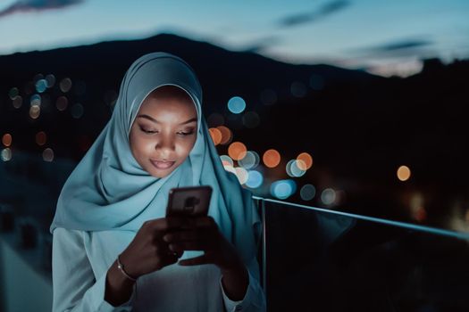 Young Muslim woman wearing scarf veil on urban city street at night texting on a smartphone with bokeh city light in the background. High-quality photo