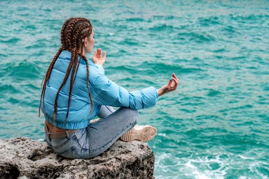 A woman in a blue jacket sits on a rock above a cliff above the sea, looking at the stormy ocean. Girl traveler rests, thinks, dreams, enjoys nature. Peace and calm landscape, windy weather