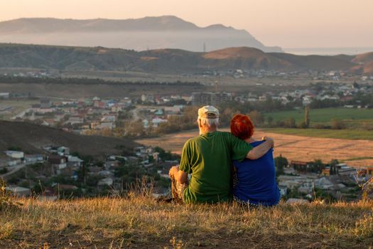 An elderly couple sits on a mountain with their backs with a beautiful view of the mountains and the sea in the distance