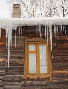 Melting transparent icicles hang on the edge of the roof. Against the background of the wooden wall of the old house. Large cascades, even beautiful rows. Cloudy winter day, soft light.