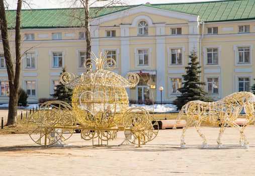 Golden street sculpture of a horse with a carriage on a frame of light bulbs. Spring, snow melts, in the background is a wall of a multi-storey building with windows. Day, cloudy weather, warm light.