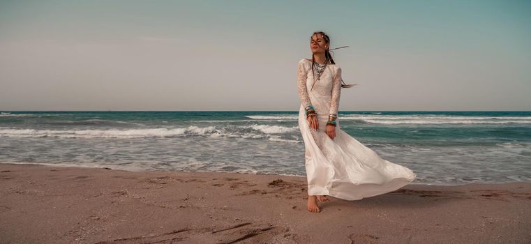 Model in boho style in a white long dress and silver jewelry on the beach. Her hair is braided, and there are many bracelets on her arms