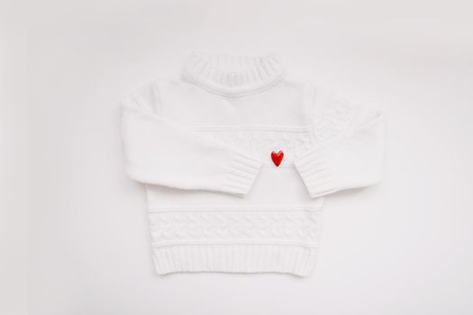 White woolen sweater with a red heart shaped pin on white background top view. Love, romance, fashion concept. Flat lay, copy space.