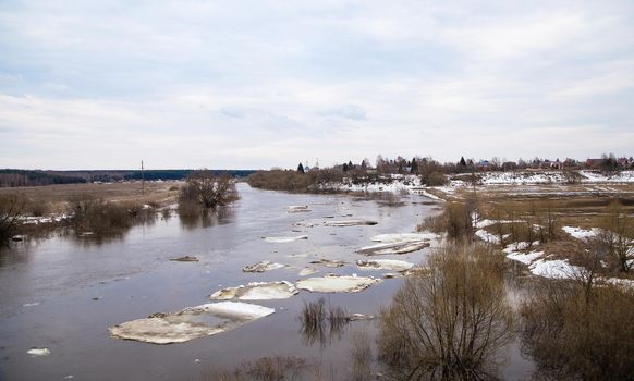 Several large dirty white ice floes are floating down the river. Spring, snow melts, dry grass all around, floods begin and the river overflows. Day, cloudy weather, soft warm light.