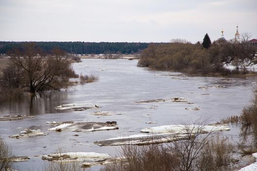 Slowly, small ice floes float down the river. Spring, snow melts, dry grass all around, floods begin and the river overflows. Day, cloudy weather, soft warm light.