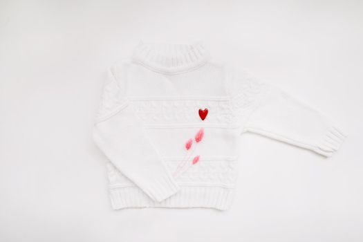 White woolen sweater with a red heart shaped pin on white background top view. Love, romance, fashion concept. Flat lay, copy space.