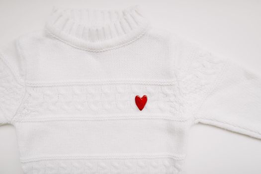 White woolen sweater with a red heart shaped pin on white background top view. Love, romance, fashion concept. Flat lay, copy space.