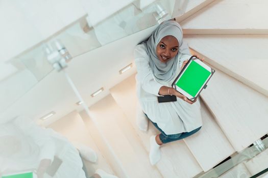 Young African modern Muslim woman using tablet computer while sitting on the stairs at home wearing hijab clothes top view. High-quality photo