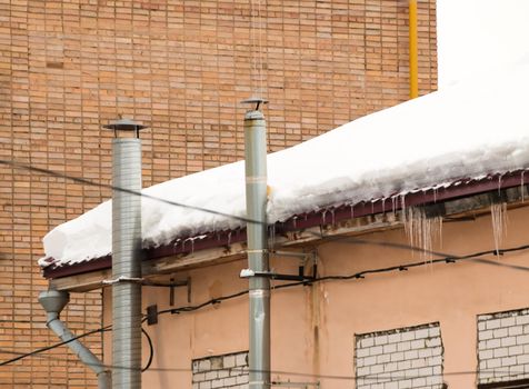 Two high metal chimneys on the roof of the house. There is white snow on the surface. Against the background of a gray sky. Cloudy winter day, soft light.