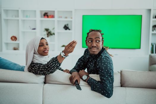 African Couple Sitting On Sofa Watching TV Together Chroma Green Screen Woman Wearing Islamic Hijab Clothes. High quality photo