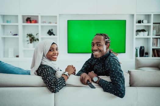 African Couple Sitting On Sofa Watching TV Together Chroma Green Screen Woman Wearing Islamic Hijab Clothes. High quality photo