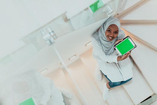 Young African modern Muslim woman using tablet computer while sitting on the stairs at home wearing hijab clothes top view. High-quality photo