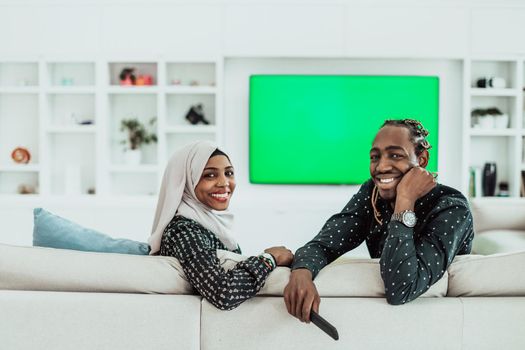 African Couple Sitting On Sofa Watching TV Together Chroma Green Screen Woman Wearing Islamic Hijab Clothes. High quality photo