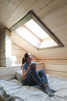 Young woman drinking tea or coffee in bed and looking out the window in the morning at home.