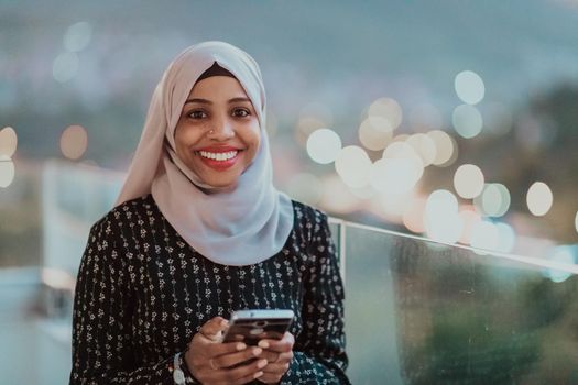 Young Muslim woman wearing scarf veil on urban city street at night texting on a smartphone with bokeh city light in the background. High-quality photo