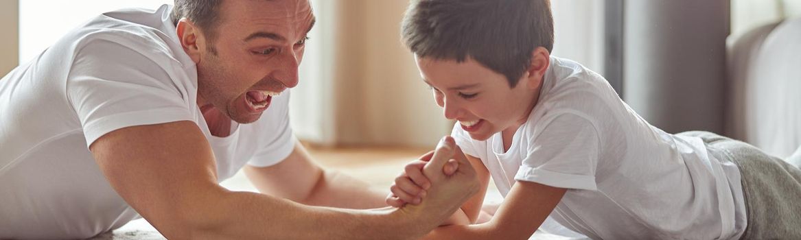 Jolly father is having fun with little boy on floor in living room and doing armwrestling battle