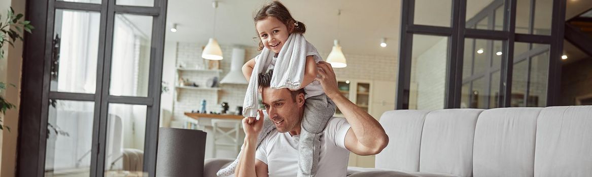 Cheerful man is holding girl on shoulders while sitting on floor in living room after workout indoors